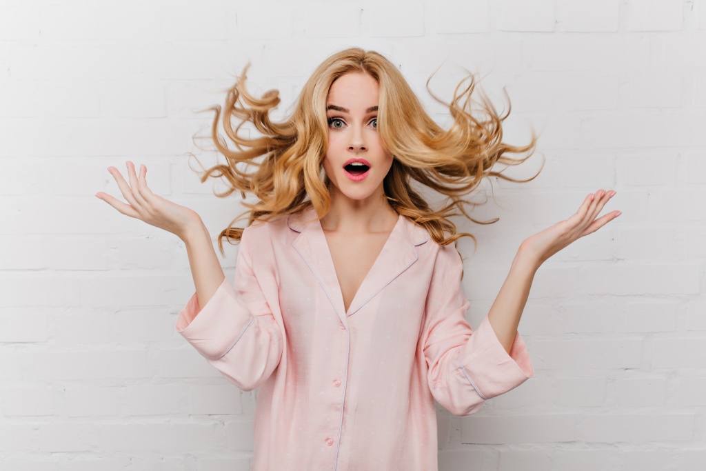 ecstatic-blue-eyed-woman-with-long-blonde-hair-posing-in-front-of-white-bricked-wall-indoor-shot-of-surprised-girl-in-beautiful-pink-pyjamas.jpg